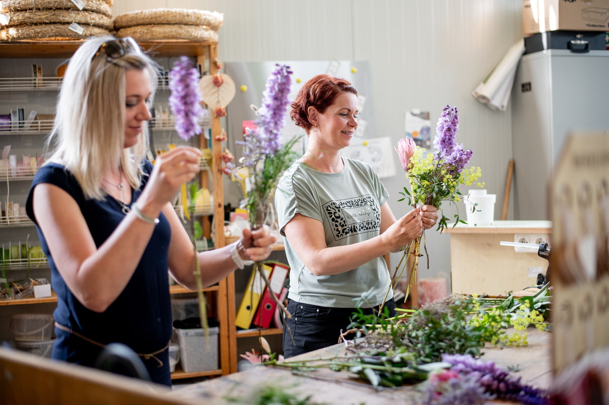 Zwei Floristinnen arrangieGartenarbeiter arrangiert Pflanzen und Produkte auf Verkaufstischen im Gartencenterren frische Blumen in einer Werkstatt; im Vordergrund bindet eine Frau ein Bouquet, während im Hintergrund weitere Blumen vorbereitet werden.