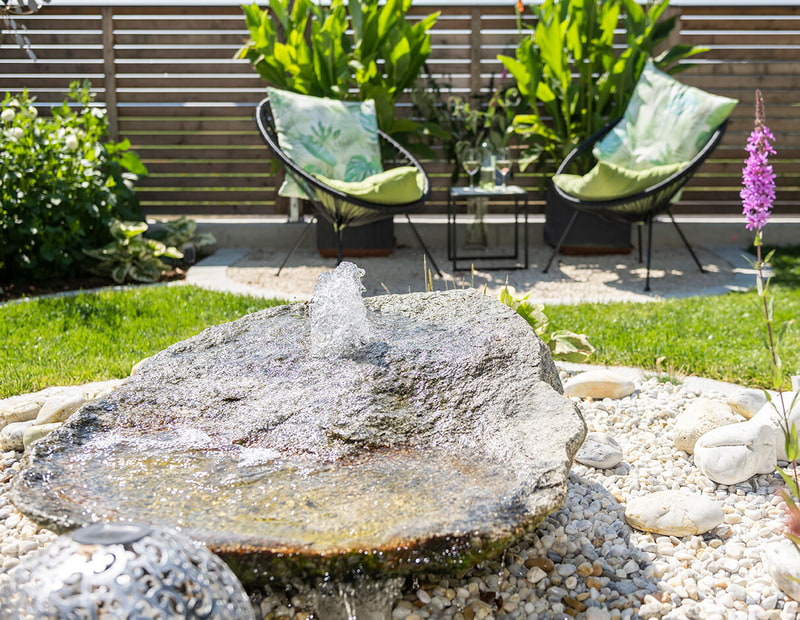 Naturstein-Wasserspiel auf Rasen mit Loungesessel und Holzsichtschutz im Hintergrund