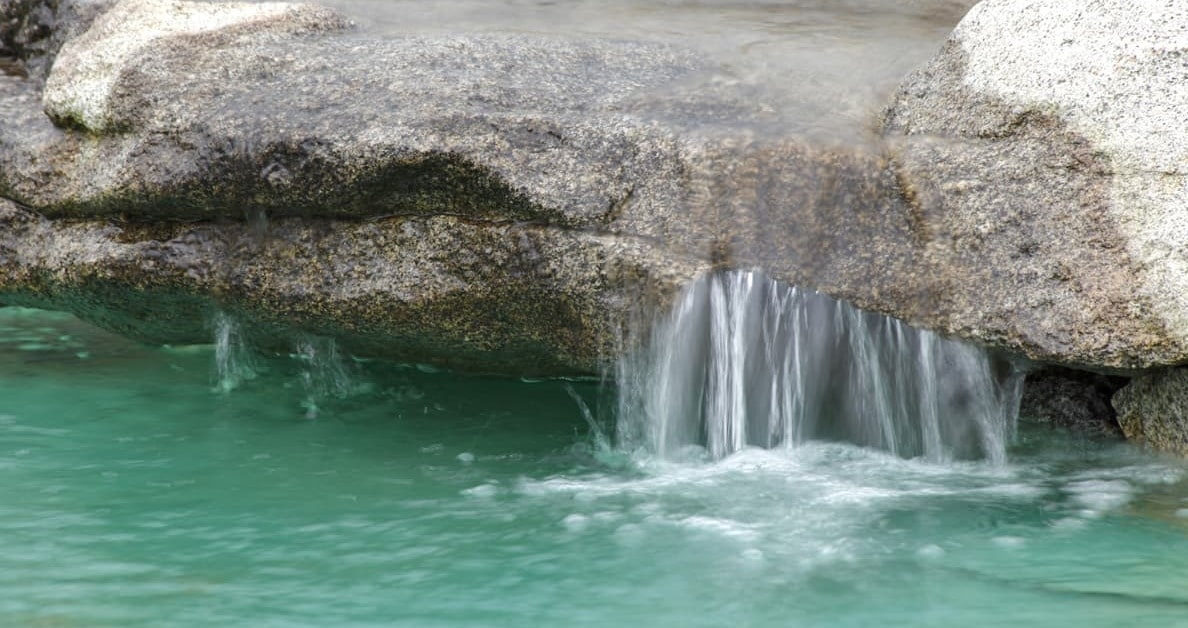 Natürlich gestalteter Wasserfall aus großen Natursteinen, bei dem klares Wasser in einen grünen Gartenpool fließt